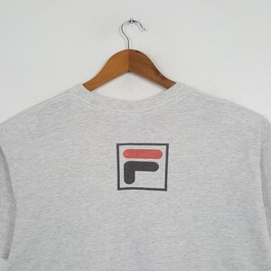 May include: A light gray sweatshirt with a black, red, and white logo. The logo is a stylized "F" and "I" inside a black square. The sweatshirt is hanging on a wooden hanger.