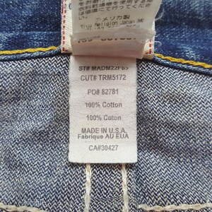 May include: Close-up of a denim garment tag. The tag displays text including "ST# MADM22F83", "CUT# TRM5172", "PO# 82781", and "100% Cotton". The tag also includes "MADE IN U.S.A." and "Fabrique AU EUA". The background is the blue denim fabric.