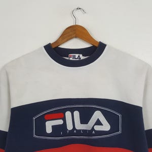 May include: A vintage Fila sweatshirt in white, navy blue, and red. The sweatshirt features the Fila logo in white with the word "ITALIA" underneath. The collar and cuffs are navy blue. The sweatshirt is hanging on a wooden hanger.