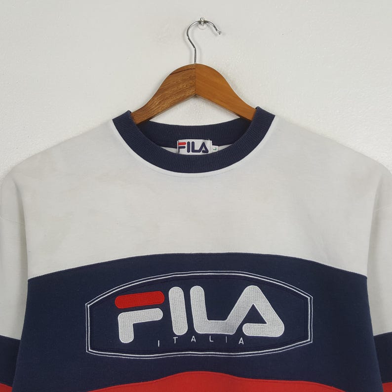 May include: A vintage Fila sweatshirt in white, navy blue, and red. The sweatshirt features the Fila logo in white with the word "ITALIA" underneath. The collar and cuffs are navy blue. The sweatshirt is hanging on a wooden hanger.