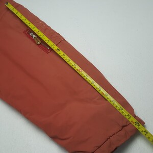 May include: Rust-colored jacket with a zippered pocket and a yellow measuring tape laid across it. The tape shows measurements in feet and inches. The jacket's fabric appears smooth and slightly shiny.