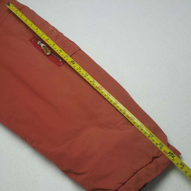 May include: Rust-colored jacket with a zippered pocket and a yellow measuring tape laid across it. The tape shows measurements in feet and inches. The jacket's fabric appears smooth and slightly shiny.
