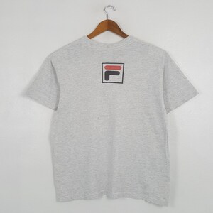 May include: A light gray t-shirt with a black, red, and white logo on the back. The logo is a square with a black border, containing a red horizontal bar and a black 'L' shape. The shirt is hanging on a wooden hanger.