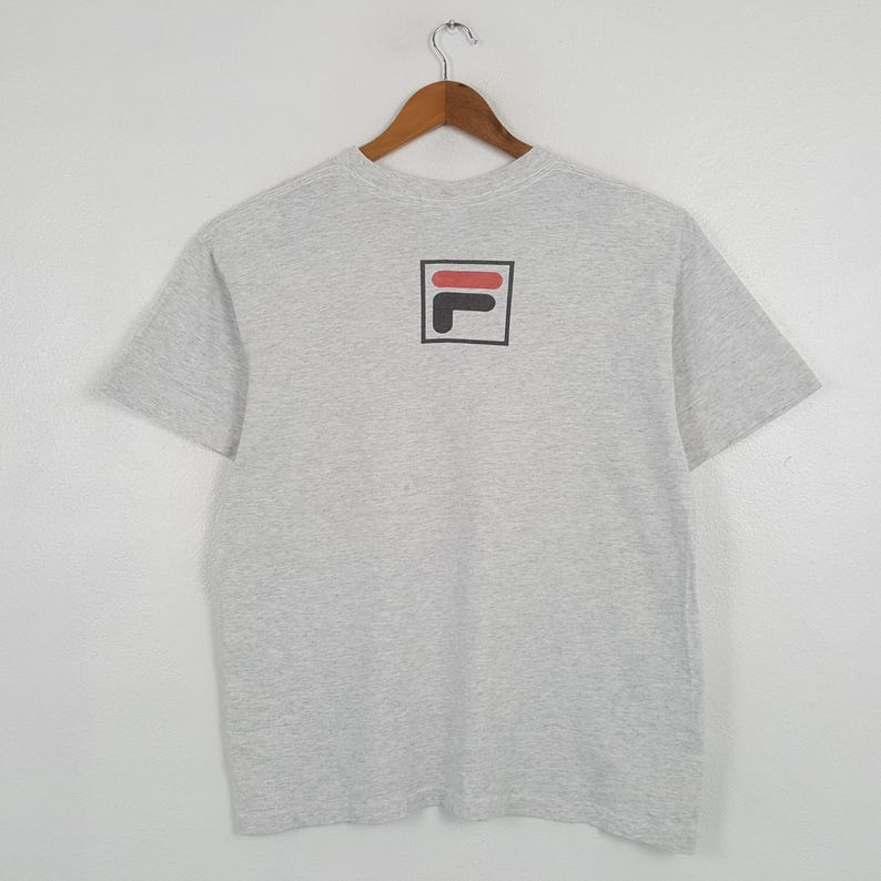 May include: A light gray t-shirt with a black, red, and white logo on the back. The logo is a square with a black border, containing a red horizontal bar and a black 'L' shape. The shirt is hanging on a wooden hanger.