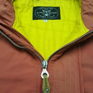 May include: Close-up of a rust-colored jacket with a yellow lining. The jacket features a full zipper and a label that reads "Avirex Officer's O'Coat Field." The jacket is a size medium.