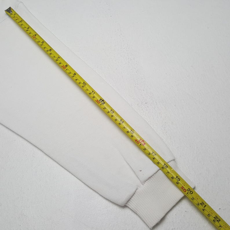 May include: A white long-sleeved sweatshirt sleeve with a ribbed cuff. A yellow measuring tape is laid across the sleeve, showing measurements in inches.