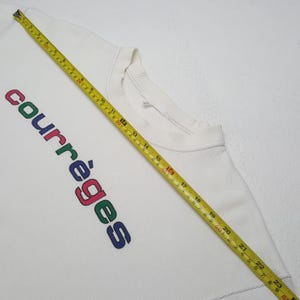 May include: White sweatshirt with the word "courr&egrave;ges" vertically printed in colorful letters. The sweatshirt has a ribbed crew neck and a ribbed hem. A yellow measuring tape is laid across the sweatshirt.