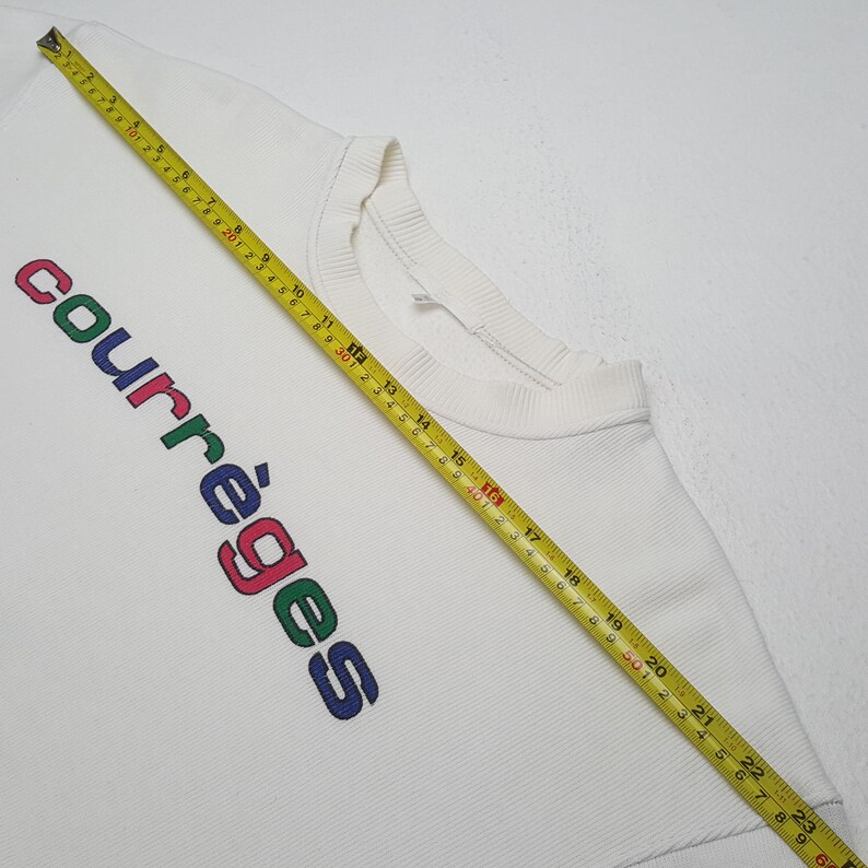 May include: White sweatshirt with the word "courr&egrave;ges" vertically printed in colorful letters. The sweatshirt has a ribbed crew neck and a ribbed hem. A yellow measuring tape is laid across the sweatshirt.