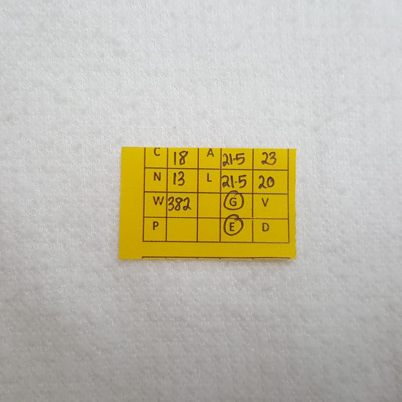 May include: A small, yellow rectangular tag with handwritten black text. The tag contains several rows and columns of numbers and letters, including "C 18", "N 13", and "W 382". The tag is placed on a white, textured surface.