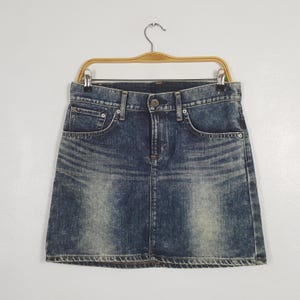 May include: A blue denim skirt with a classic five-pocket design, button closure, and a faded wash. The skirt is hanging on a wooden hanger against a white background. The skirt has a straight hemline.