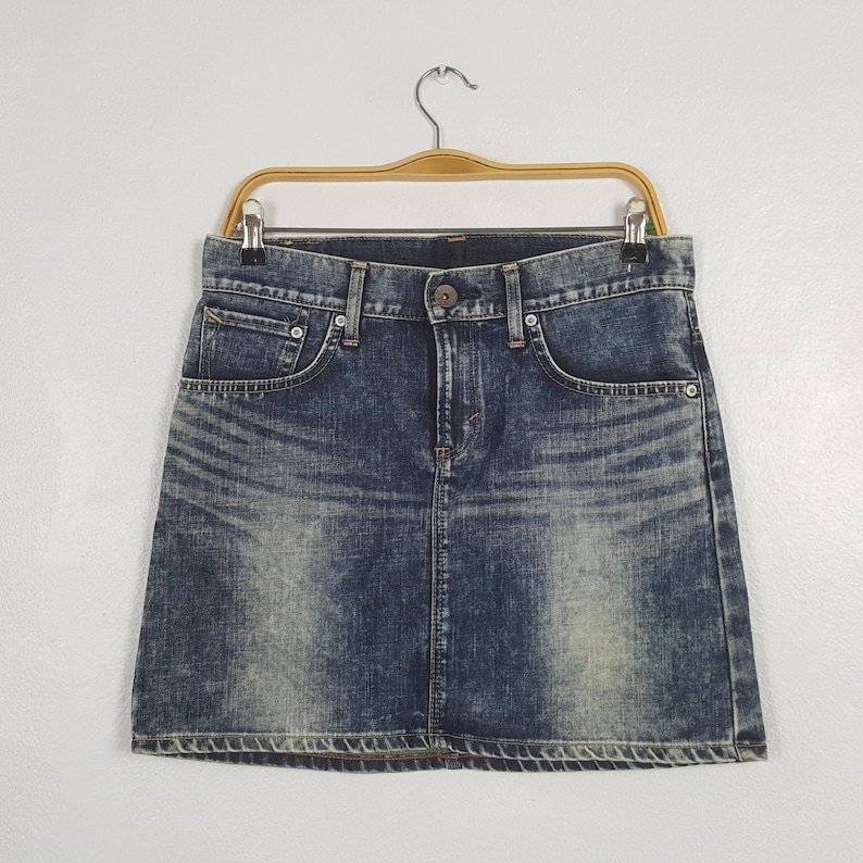 May include: A blue denim skirt with a classic five-pocket design, button closure, and a faded wash. The skirt is hanging on a wooden hanger against a white background. The skirt has a straight hemline.