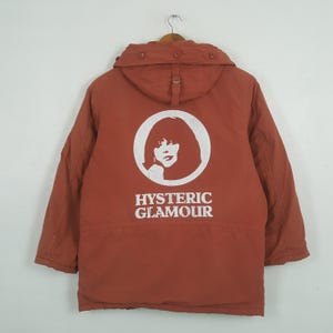 May include: A rust-colored jacket with a hood, hanging on a wooden hanger. The back of the jacket features a white graphic of a woman's face inside a circle, with the words "HYSTERIC GLAMOUR" below.