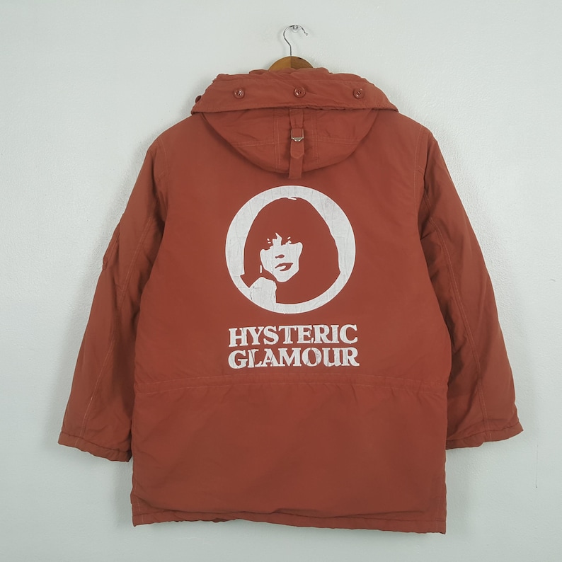 May include: A rust-colored jacket with a hood, hanging on a wooden hanger. The back of the jacket features a white graphic of a woman's face inside a circle, with the words "HYSTERIC GLAMOUR" below.
