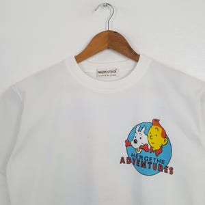 May include: White crew neck t-shirt with a graphic of two cartoon characters in a blue circle. The text "HERGE THE ADVENTURES" is below the graphic. The shirt is hanging on a wooden hanger.