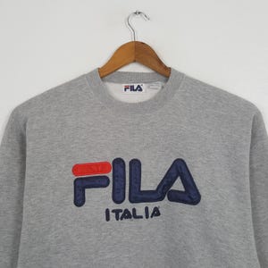 May include: A gray crew neck sweatshirt with the FILA logo in red and navy blue. The word "ITALIA" is embroidered below the logo. The sweatshirt is hanging on a wooden hanger.