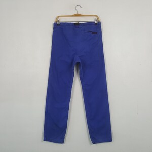 May include: Bright blue pants hanging on a wooden hanger. The pants have a straight leg design and a small pocket on the back. The fabric appears to be a durable cotton or blend. The pants are a solid, vibrant color.