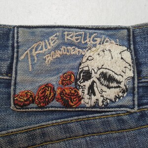 May include: Close-up of a denim patch with embroidered details. The patch features the text "TRUE RELIGION BRAND JEANS" in cream-colored thread. A skull and red roses are also embroidered on the patch.
