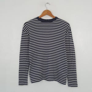 May include: A long-sleeved shirt with horizontal navy blue and white stripes. The shirt is hanging on a wooden hanger against a plain white background. The shirt has a crew neck and a relaxed fit.