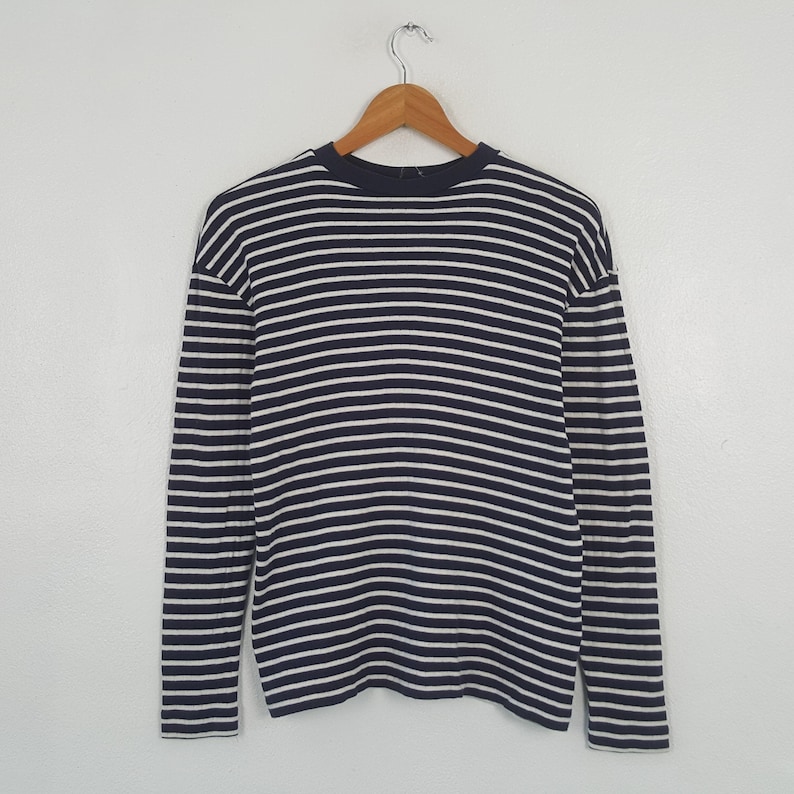 May include: A long-sleeved shirt with horizontal navy blue and white stripes. The shirt is hanging on a wooden hanger against a plain white background. The shirt has a crew neck and a relaxed fit.