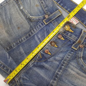 May include: Close-up of blue denim jeans with a button fly and yellow stitching. A yellow measuring tape shows a measurement of approximately 15 inches. The jeans have a distressed look with faded areas and brown spots.