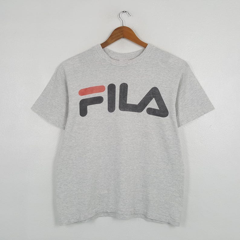 May include: A light gray t-shirt with the word "FILA" printed across the chest in a bold, black font with a red accent. The shirt is short-sleeved and hangs from a wooden hanger against a plain white background.
