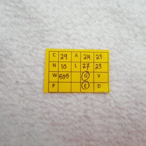 May include: A small, yellow rectangular tag with black handwritten text and numbers. The tag is divided into several boxes, each containing a letter or number. The tag is on a white, textured surface.