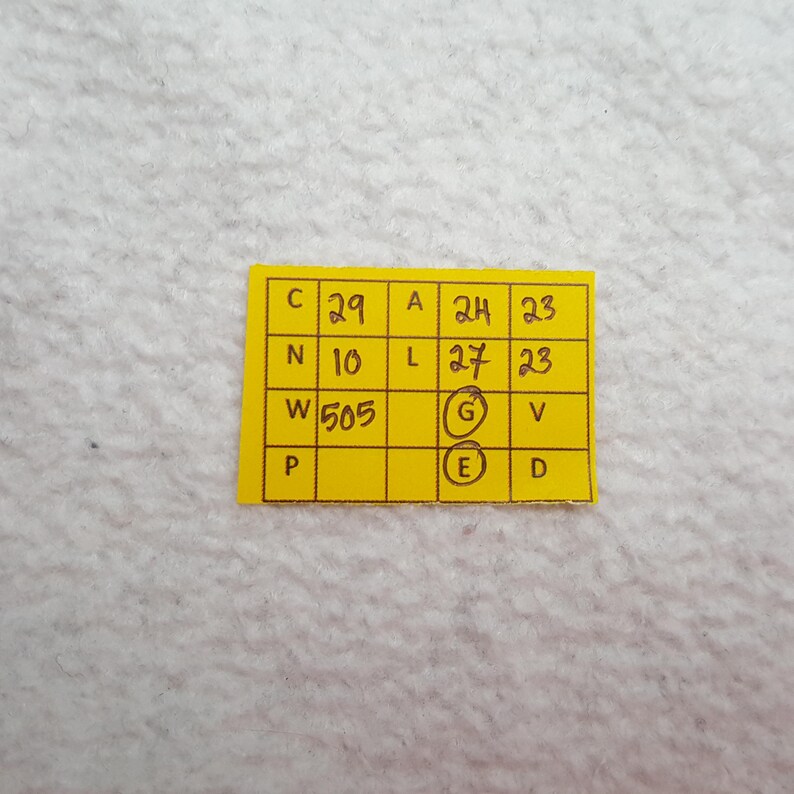 May include: A small, yellow rectangular tag with black handwritten text and numbers. The tag is divided into several boxes, each containing a letter or number. The tag is on a white, textured surface.