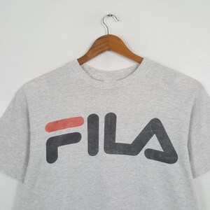 May include: A light gray t-shirt with the FILA logo in black and red. The shirt is hanging on a wooden hanger against a white background. The logo is large and centered on the front of the shirt.