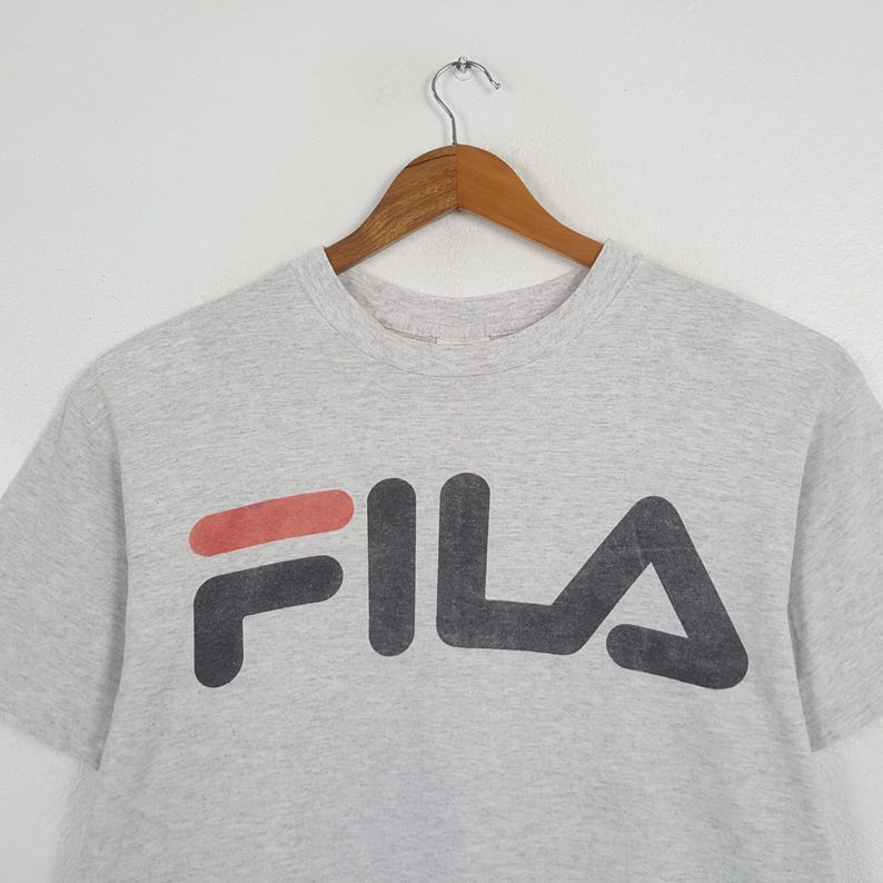 May include: A light gray t-shirt with the FILA logo in black and red. The shirt is hanging on a wooden hanger against a white background. The logo is large and centered on the front of the shirt.
