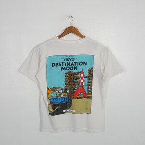 May include: White t-shirt featuring a colorful graphic from "The Adventures of Tintin: Destination Moon." The design shows a rocket, a blue truck, and the text "DESTINATION MOON."
