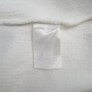 May include: Close-up of a white fabric with a sewn-in label. The label is white and rectangular, with text and symbols printed on it. The fabric has a textured appearance.