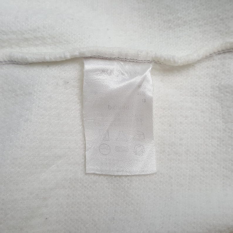 May include: Close-up of a white fabric with a sewn-in label. The label is white and rectangular, with text and symbols printed on it. The fabric has a textured appearance.