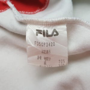 May include: Close-up of a white garment tag with the FILA logo in gray. The tag includes the text "FD5GF3420" and Japanese characters. The fabric composition is listed as 70% cotton and 30% polyester.