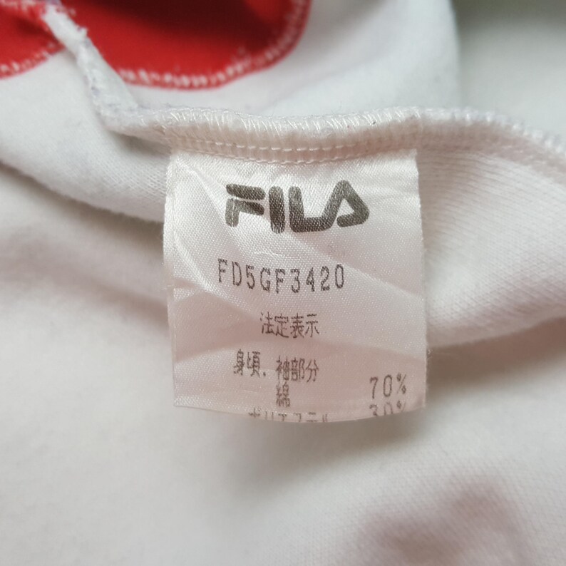 May include: Close-up of a white garment tag with the FILA logo in gray. The tag includes the text "FD5GF3420" and Japanese characters. The fabric composition is listed as 70% cotton and 30% polyester.