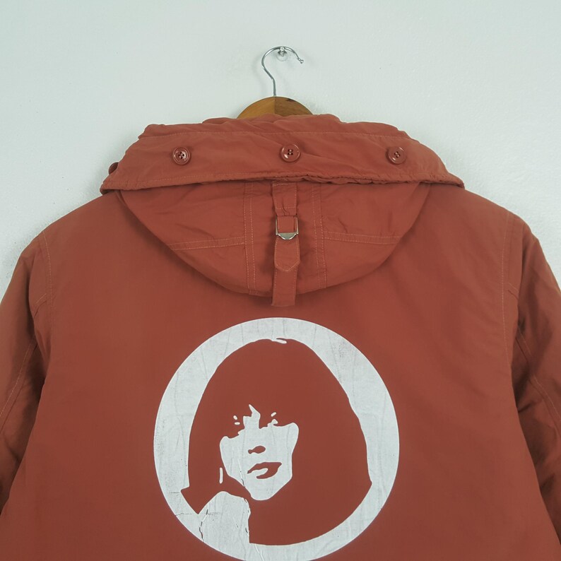 May include: Rust-colored jacket with a hood and a white graphic of a woman's face inside a circle on the back. The jacket has a buckle closure and buttons on the hood.