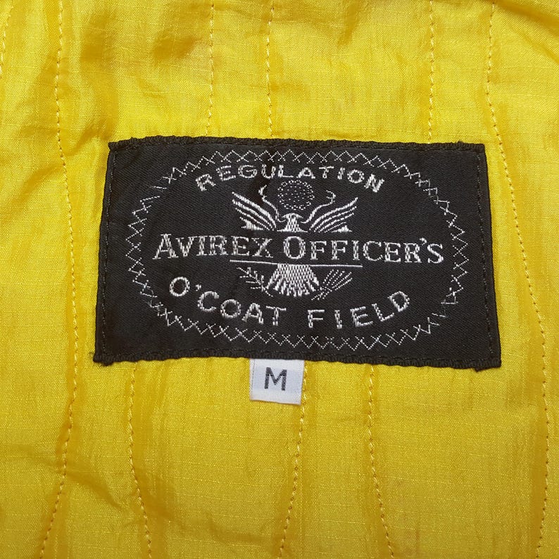 May include: Close-up of a yellow quilted fabric with a black label. The label reads "REGULATION AVIREX OFFICER'S O'COAT FIELD" with an eagle emblem. A white tag with the letter "M" is below the label.