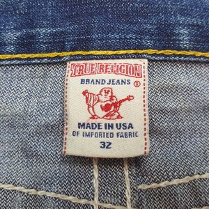 May include: Close-up of a True Religion brand jeans tag. The tag is white with red text and a red graphic of a Buddha playing a guitar. The text reads "TRUE RELIGION BRAND JEANS", "MADE IN USA OF IMPORTED FABRIC", and "32".