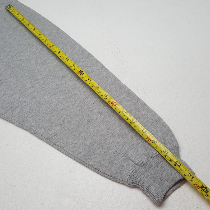 May include: A gray sweatshirt sleeve with a yellow measuring tape laid across it. The tape shows measurements in inches, with the sleeve's cuff at the 24-inch mark. The fabric is a solid, light gray color.