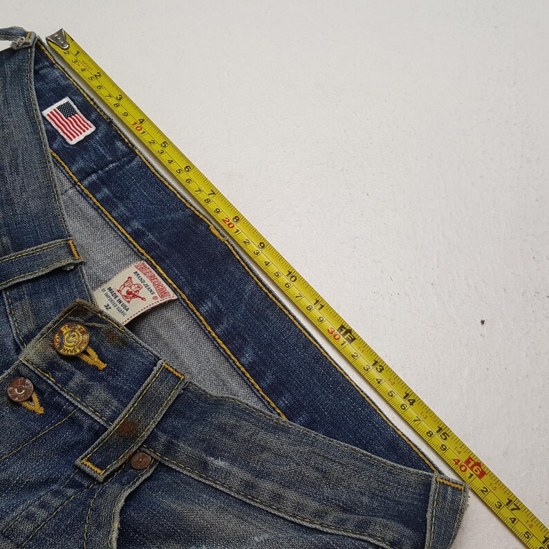 May include: Close-up of blue denim jeans with a yellow measuring tape. The jeans have a button closure and a small American flag patch. The tag reads "MADE IN USA". The jeans measure approximately 17 inches.