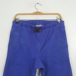 May include: Bright blue pants with an elastic waistband and drawstring, hanging on a wooden hanger. The pants have a relaxed fit and are made of a solid-colored fabric. The brand name is visible on the waistband.
