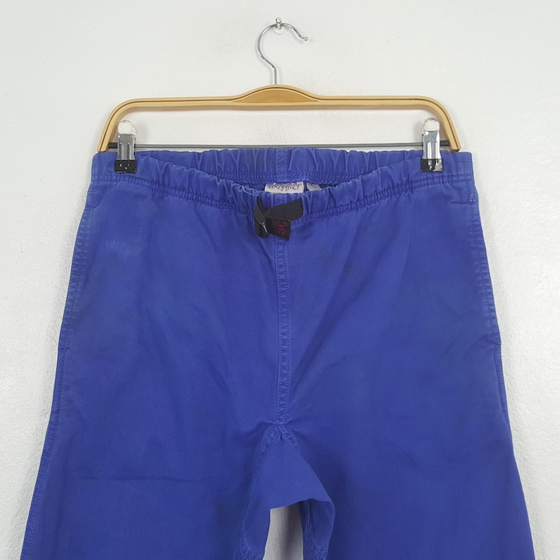 May include: Bright blue pants with an elastic waistband and drawstring, hanging on a wooden hanger. The pants have a relaxed fit and are made of a solid-colored fabric. The brand name is visible on the waistband.