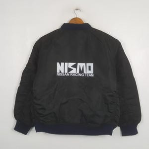 Può includere: Un giubbotto bomber nero con colletto, polsini e orlo a coste blu scuro. Il retro presenta la scritta bianca NISMO e NISSAN RACING TEAM stampata al centro. La giacca è realizzata in un tessuto nero liscio e a tinta unita.