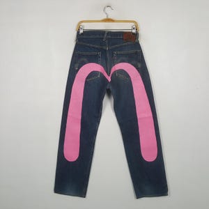 Puede incluir: Vaqueros de mezclilla azul oscuro con un gran gráfico de gaviota pintado en rosa brillante en los bolsillos traseros. Los pantalones tienen un diseño clásico de cinco bolsillos, un parche de cuero marrón en la cintura y un gancho metálico.