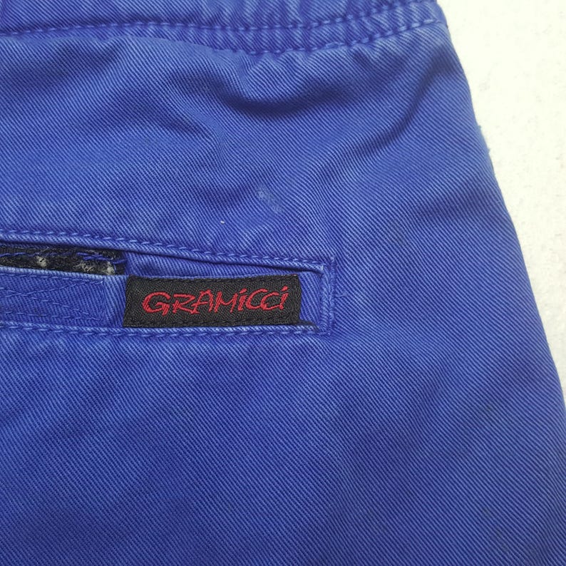 May include: Close-up of blue Gramicci shorts. The shorts are a solid blue color with a small red and black Gramicci label sewn on the back pocket. The fabric has a textured appearance.