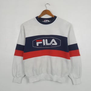 May include: White Fila sweatshirt with navy blue and red stripes across the chest. The Fila logo is prominently displayed in white within a navy blue rectangle. The sweatshirt has a crew neck and long sleeves.