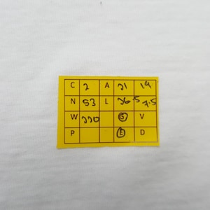 May include: A small, yellow rectangular tag with black handwritten text and numbers. The tag is divided into a grid of boxes, each containing a letter or number. The tag is placed on a white background.