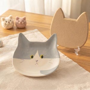 Può includere: Un piatto in ceramica a forma di gatto con muso grigio e bianco, occhi gialli e un piccolo naso rosa. Accanto si trova un sottopentola in sughero color marrone chiaro a forma di gatto. Sullo sfondo, due piccoli ornamenti in lana a forma di gatto.