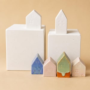 Pode incluir: Duas figuras de casa de cerâmica branca assentam sobre dois blocos retangulares brancos. Abaixo estão quatro figuras de casa de cerâmica mais pequenas em azul, rosa claro, verde com base laranja e rosa claro, com detalhes de janelas.
