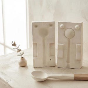 Può includere: Due stampi rettangolari in gesso bianco sporco con forme di cucchiaio incavate, posizionati in verticale. Un piccolo vaso rotondo in ceramica bianco sporco contiene un rametto di eucalipto. Un cucchiaio finito con coppa bianca e manico in legno è appoggiato davanti.