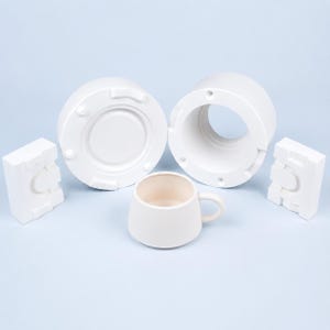 May include: A white ceramic mug positioned in front of a multi-part plaster slip casting mold. The mold consists of a circular base piece, a cylindrical main body section, and two rectangular side components, all featuring a smooth matte finish.